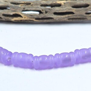 May include: A strand of light purple beads with a unique, rounded shape. The beads are strung together with a silver-colored wire. The background is white with a piece of driftwood.