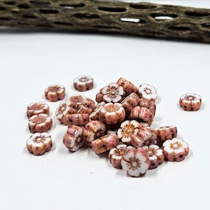 May include: A collection of flower-shaped beads in shades of pink, white, and brown. The beads have a glossy finish and are scattered on a white surface. The beads are likely for jewelry making or other craft projects.