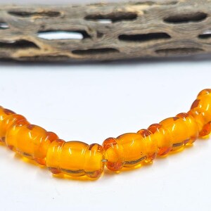 May include: A strand of amber-colored beads, arranged in a gentle curve, against a white background. The beads are cylindrical with rounded ends. A piece of weathered wood is in the background.