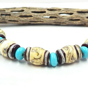 May include: A necklace featuring cream-colored, barrel-shaped beads with brown swirls, separated by turquoise blue beads. The beads are strung together, creating a unique jewelry piece. A piece of driftwood is in the background.