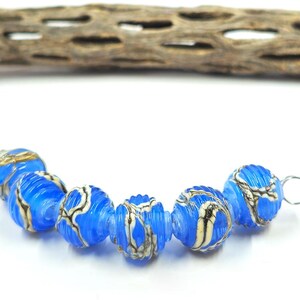 May include: A strand of vibrant blue glass beads with a ribbed texture and intricate white and black patterns. Each bead is round, with a silver loop for stringing. The beads are displayed against a white background, with a piece of wood in the background.