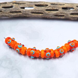 May include: A strand of orange glass beads with blue and red accents. The beads have a rounded shape with a textured surface. The beads are strung on a silver wire and are arranged in a curved line. The background includes a piece of driftwood.