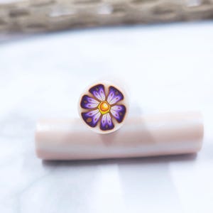 Puede incluir: Una caña de arcilla polimérica con un diseño floral. La flor tiene pétalos morados con un centro amarillo y está sobre un fondo blanco. La caña es cilíndrica y de color rosa claro.