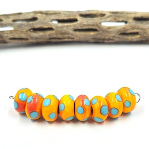 May include: A strand of colorful glass beads. The beads are oval-shaped, with a vibrant orange and yellow base color, and adorned with light blue polka dots. A piece of textured wood is in the background.
