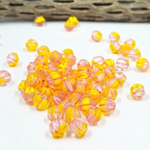 Peut inclure: Un ensemble de perles rondes translucides avec un motif rayé jaune et rose. Les perles sont dispersées sur une surface blanche, avec un morceau de bois flotté en arrière-plan. Ces perles sont pour la création de bijoux.