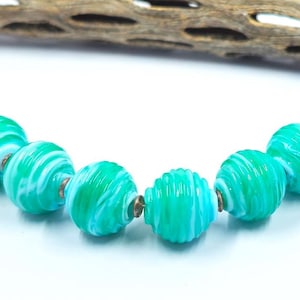 May include: A strand of six round, ribbed glass beads featuring a marbled pattern of bright turquoise, emerald green, and white swirls. Small, metallic-toned spacer beads are positioned between each of the larger glass beads along the string.