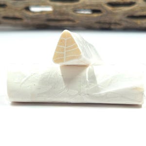 May include: A cylindrical object wrapped in translucent white paper with a visible leaf-like vein pattern. A second, smaller triangular-shaped piece with a similar light-colored organic texture and vein pattern rests on top of the cylindrical base.