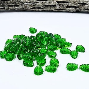 Può includere: Una collezione di perline a forma di foglia di vetro verde traslucido sparse su una superficie bianca. Le perline, con dettagliati motivi a venature di foglie, variano per dimensioni e forma. Un pezzo di legno strutturato è sullo sfondo.