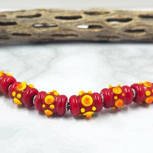 May include: A strand of red glass beads with yellow and orange dot patterns. The beads are strung together with silver-colored metal spacers. A piece of dried wood is in the background.