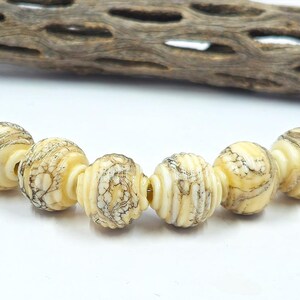 May include: A strand of six cream-colored, round beads with a marbled, brown pattern. Each bead has a ribbed texture and is connected with a silver-colored metal loop. A piece of weathered wood is in the background.