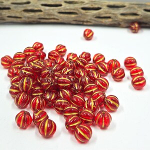 May include: A collection of red and gold striped beads. The beads are round with a ribbed texture and a transparent, glossy finish. The gold stripes run horizontally around the beads, creating a decorative pattern. These beads are suitable for jewelry making.