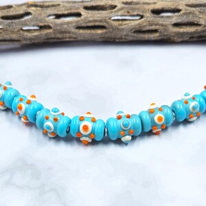 May include: A strand of turquoise blue glass beads with orange and white accents. The beads are cylindrical with raised circular designs. The beads are strung together on a silver-colored wire. The beads are likely for jewelry making.