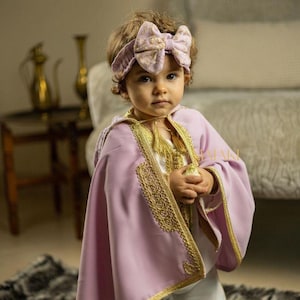 May include: A young child wearing a lavender robe with gold embroidery and a floral headband. The robe has a tassel and is worn over a white shirt.