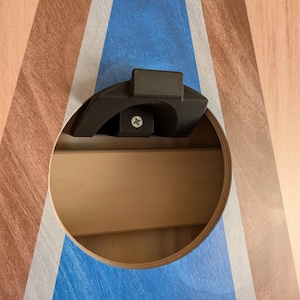 May include: Close-up of a cornhole board with a circular hole and a black plastic handle. The board features a blue and white design on a wood-grain background. A screw is visible inside the handle.