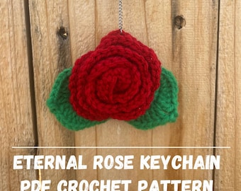 Peut inclure: Un porte-clés fait à la main avec une rose rouge et des feuilles vertes. Il est suspendu à une chaîne argentée. Le texte "ETERNAL ROSE KEYCHAIN PDF CROCHET PATTERN" est affiché en dessous. Fond en bois.