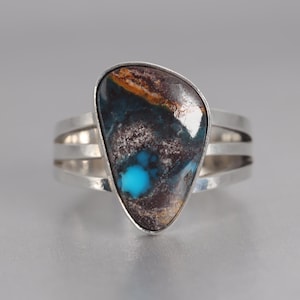 Könnte beinhalten: Ein Silberring mit einem großen, unregelmäßig geformten Stein. Der Stein weist eine Mischung aus Türkis, Braun und Schwarz auf. Das Ringband hat ein dreifaches Design.