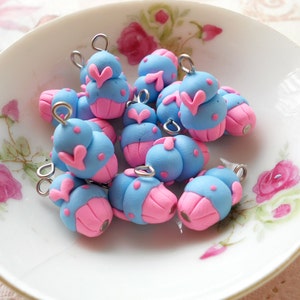 Puede incluir: Una colección de abalorios de arcilla polimérica hechos a mano en un plato blanco con estampado floral. Cada abalorio es un diseño caprichoso, con una parte superior azul claro y una base rosa, con detalles de corazón y puntos rosas. Se adjuntan bucles de metal plateado.