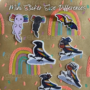 Puede incluir: Una colección de pegatinas coloridas con un ajolote, un murciélago y cuervos con detalles de arcoíris. Las pegatinas se muestran sobre un fondo marrón con diseños de arcoíris y el texto "Mini Sticker Size Differences". El nombre y el lema de la tienda también son visibles.