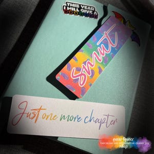 May include: A light teal notebook with a bookmark that says "Smut" in pink script over a rainbow background with eggplant illustrations. Another bookmark reads "Just one more chapter" in rainbow colors. A sticker says "This year I will give a..."
