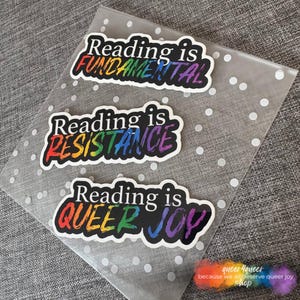Könnte beinhalten: Drei Aufkleber mit den Texten "Reading is FUNDAMENTAL", "Reading is RESISTANCE" und "Reading is QUEER JOY". Jeder Aufkleber hat einen schwarzen Rand und einen Text in Regenbogenfarben. Die Aufkleber befinden sich auf einer transparenten, gepunkteten Oberfläche.