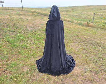 May include: A long, dark blue hooded cloak made of velvet. The cloak is floor-length and has a wide, flowing skirt. The hood is up, and the person wearing the cloak is facing away from the camera. The background is a grassy field.