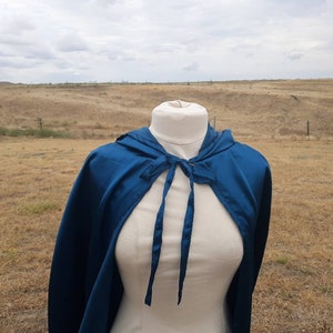 May include: A teal blue hooded cape with a tie closure. The cape is made of a soft, silky fabric and is draped over a white mannequin.