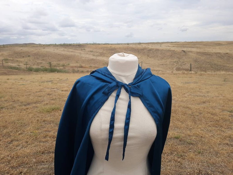 May include: A teal blue hooded cape with a tie closure. The cape is made of a soft, silky fabric and is draped over a white mannequin.