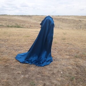 May include: A long, flowing blue cape with a hood. The cape is made of a smooth, silky fabric and is draped over a mannequin. The mannequin is standing in a field of dry grass.