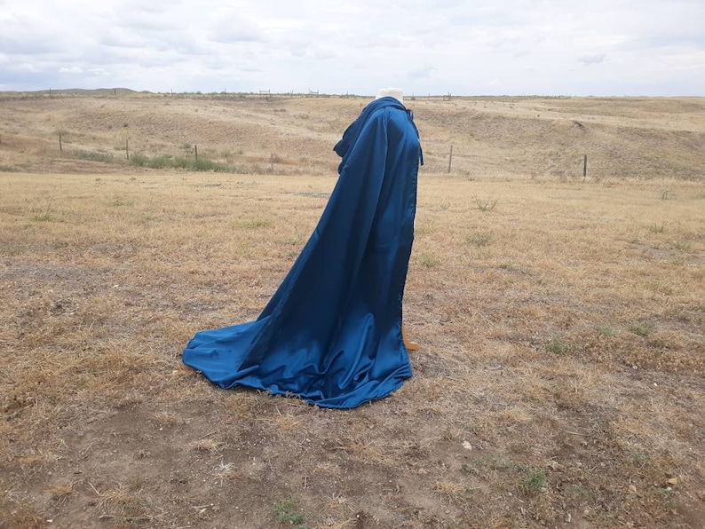 May include: A long, flowing blue cape with a hood. The cape is made of a smooth, silky fabric and is draped over a mannequin. The mannequin is standing in a field of dry grass.
