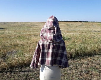May include: A hooded cape made of plaid fabric in shades of brown and white. The cape has a pointed hood and is worn on a mannequin against a backdrop of a grassy field.