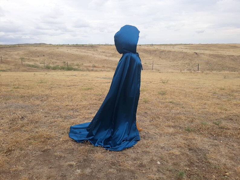 May include: A long, flowing blue satin cape with a large hood. The cape is draped over a field of dry grass.