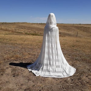 Op de afbeelding: Een witte kanten kapmantel met een lange sleep. De mantel is gefotografeerd in een veld met een blauwe lucht op de achtergrond.
