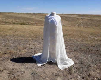 May include: A white lace cape with a hood, displayed on a mannequin against a backdrop of a grassy field.