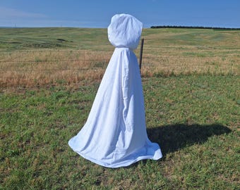 Puede incluir: Una prenda blanca con capucha drapeada sobre un poste de madera en un campo cubierto de hierba. La tela es ligera y fluida, con una capucha redondeada y una falda larga. El fondo presenta una valla, colinas y un cielo azul.