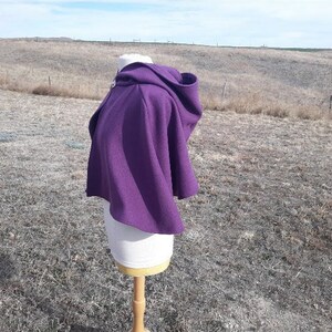 K&ouml;nnte beinhalten: Ein leuchtend lila Kapuzenponcho, der auf einer Schaufensterpuppe pr&auml;sentiert wird. Der Poncho besteht aus einem dicken, strukturierten Stoff und hat eine gro&szlig;e Kapuze und einen Verschluss am Halsausschnitt. Das Kleidungsst&uuml;ck ist zum &Uuml;berwerfen &uuml;ber die Schultern gedacht.
