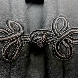 K&ouml;nnte beinhalten: Nahaufnahme eines schwarzen Kleidungsst&uuml;cks aus Stoff mit zwei dekorativen schwarzen Stoffverschl&uuml;ssen. Die Verschl&uuml;sse sind in der Mitte zu einem Knoten gebunden und haben ein Schleifendesign.