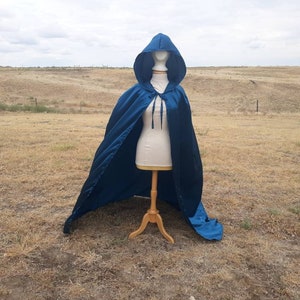 May include: A long, blue satin cape with a hood. The cape is draped over a mannequin and is displayed against a backdrop of a grassy field.