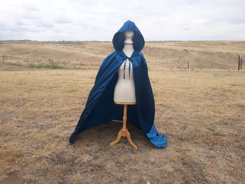 May include: A long, blue satin cape with a hood. The cape is draped over a mannequin and is displayed against a backdrop of a grassy field.