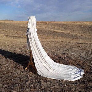 May include: A white hooded cape with a long train, displayed on a mannequin against a backdrop of a dry, grassy field.