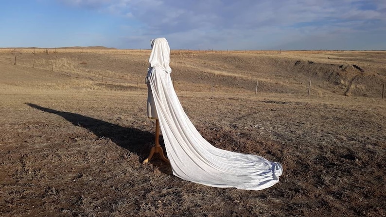 May include: A white hooded cape with a long train, displayed on a mannequin against a backdrop of a dry, grassy field.
