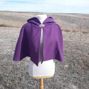 K&ouml;nnte beinhalten: Ein tiefvioletter Umhang mit Kapuze und silberner Brosche. Der Umhang wird auf einer B&uuml;ste pr&auml;sentiert, um seinen flie&szlig;enden Fall zu zeigen. Der Stoff scheint weich und strukturiert zu sein, ideal f&uuml;r Cosplay oder historische Veranstaltungen.