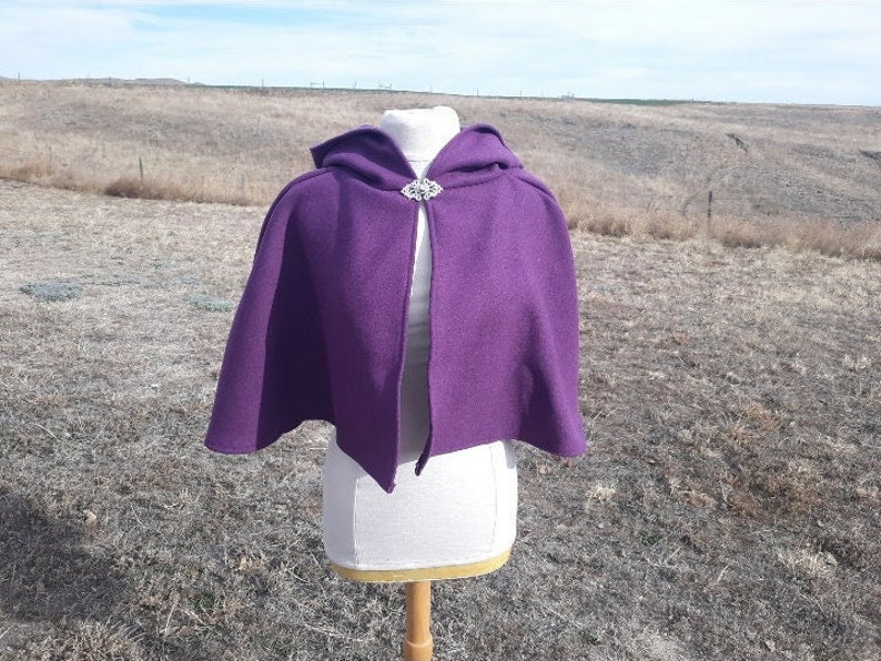 K&ouml;nnte beinhalten: Ein tiefvioletter Umhang mit Kapuze und silberner Brosche. Der Umhang wird auf einer B&uuml;ste pr&auml;sentiert, um seinen flie&szlig;enden Fall zu zeigen. Der Stoff scheint weich und strukturiert zu sein, ideal f&uuml;r Cosplay oder historische Veranstaltungen.