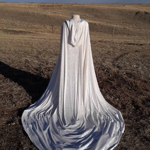 May include: A white hooded cape with a long train, made of velvet, is displayed on a mannequin in a field.