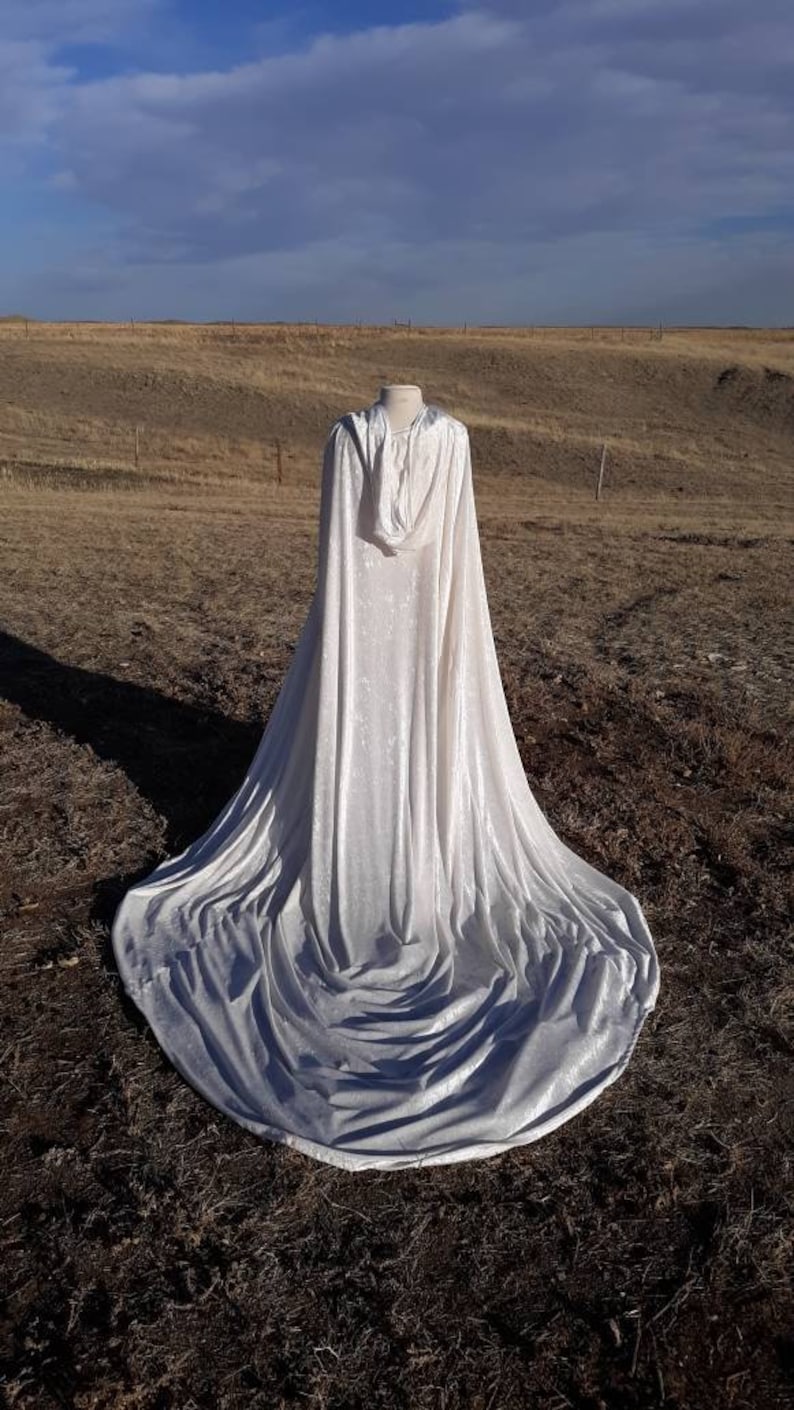 May include: A white hooded cape with a long train, made of velvet, is displayed on a mannequin in a field.
