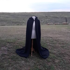 May include: A black hooded cape with a long flowing design, displayed on a mannequin against a grassy background.