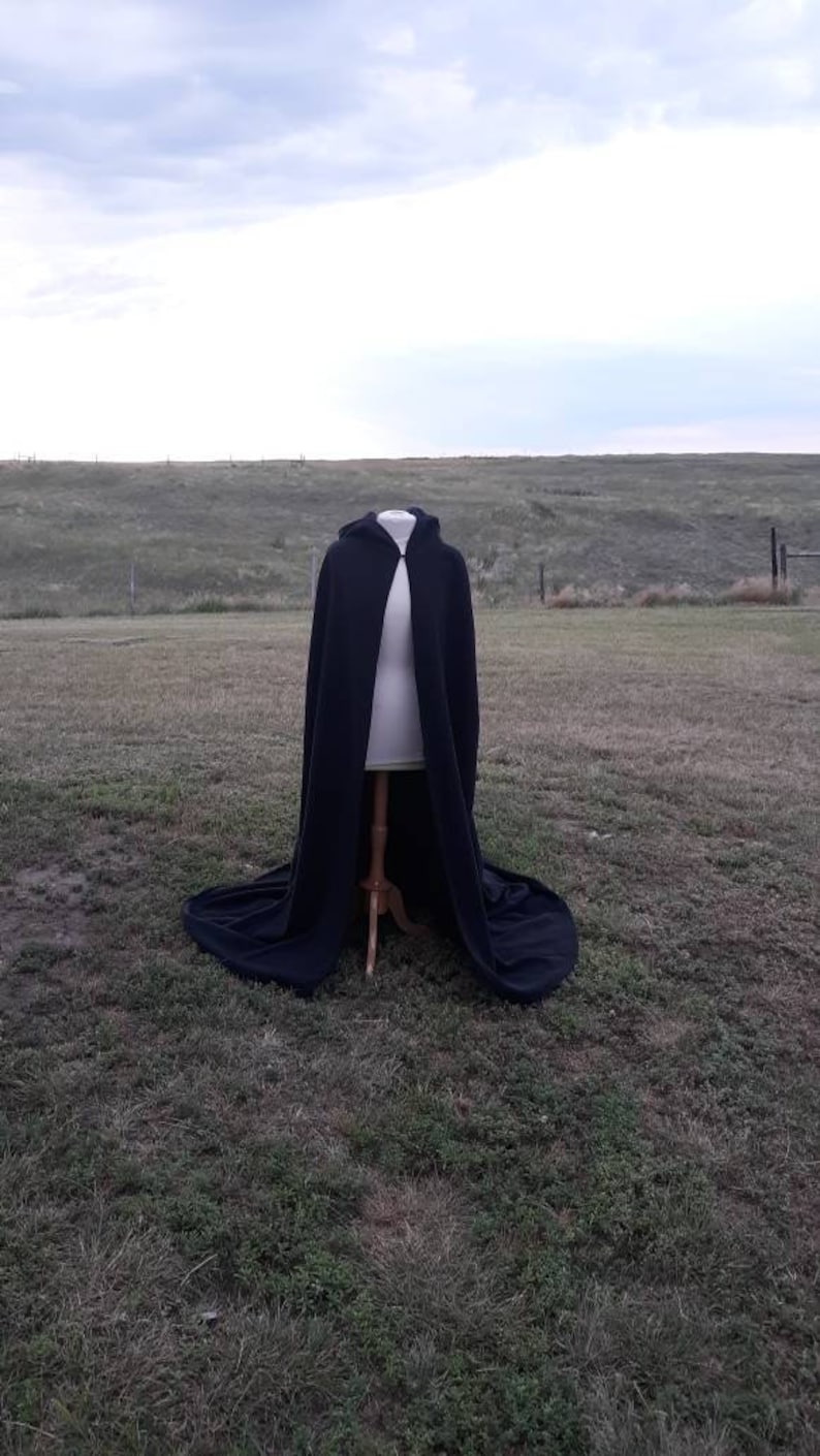 May include: A black hooded cape with a long flowing design, displayed on a mannequin against a grassy background.