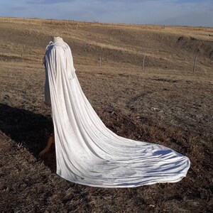 May include: A long white cape with a flowing train, displayed on a mannequin in a field.