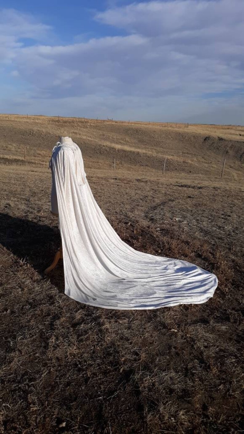 May include: A long white cape with a flowing train, displayed on a mannequin in a field.