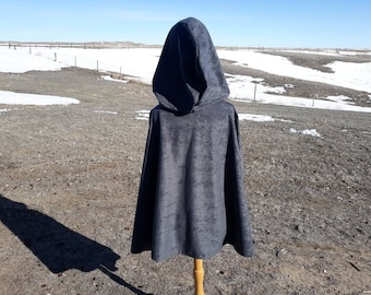 Op de afbeelding: Een donkergrijze cape met capuchon, buiten tentoongesteld op een houten standaard. De cape heeft een getextureerde uitstraling en een grote capuchon. De achtergrond toont een veld met sneeuwplekken en een heldere blauwe lucht.