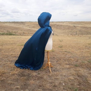 May include: A teal blue hooded cape with a satin finish. The cape is draped over a dress form and is displayed against a backdrop of a grassy field.